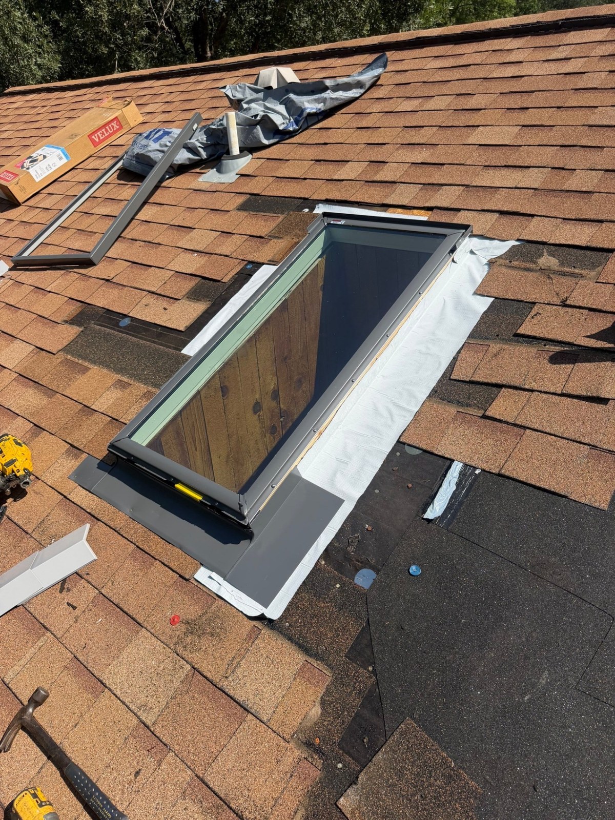 A new skylight during installation, with white waterproof membrane around the opening and gray flashing being installed, shingles cut back around it.