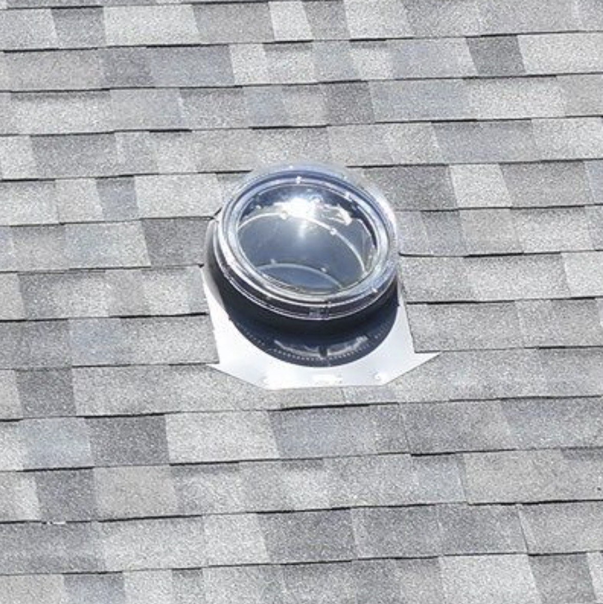Close-up of a clear solar tube dome on its metal flashing, set into gray shingles.
