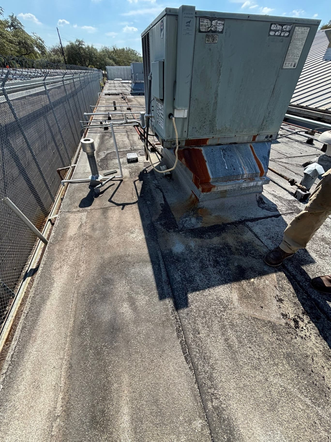 An old granulated ModBit roof around a rooftop AC unit, with black roof cement patches at its base and rust running down the curb.