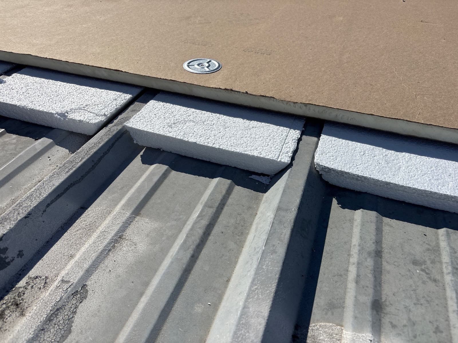 Foam strips filling the channels of an old metal roof, with insulation board fastened on top using screws and metal plates.
